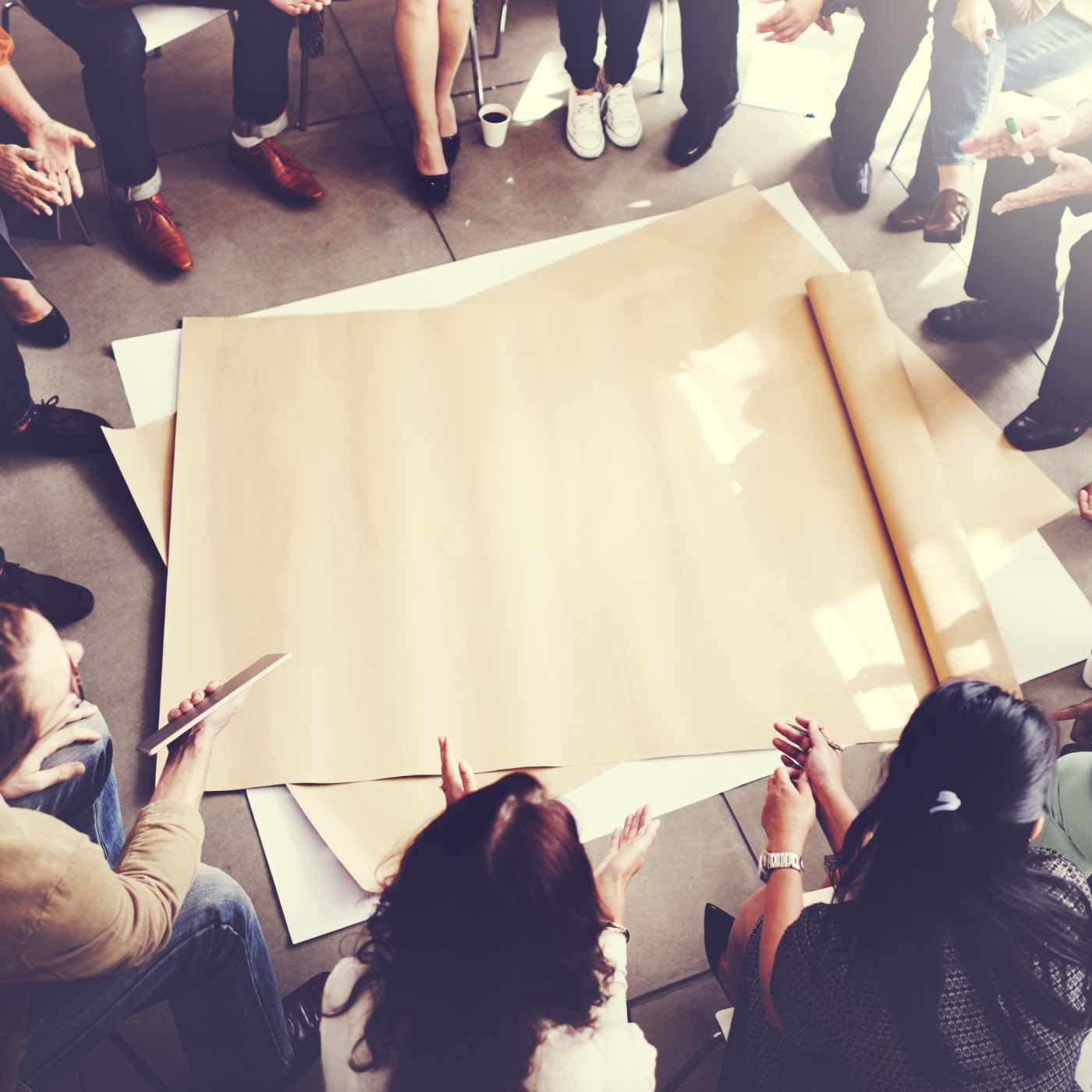 Eine Gruppe von Menschen sitzt im Kreis auf dem Boden. In der Mitte liegt ein großes Stück braunes Papier. Die Personen sprechen miteinander und gestikulieren, als würden sie gemeinsam an einer Idee oder einem Projekt arbeiten.