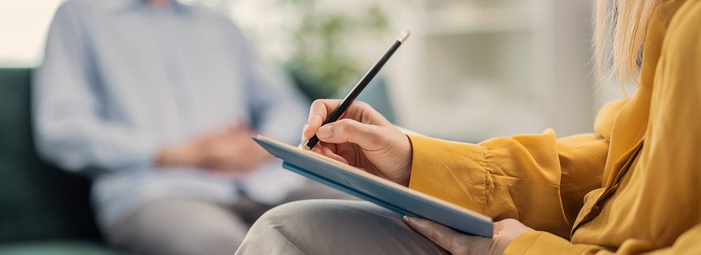 Zwei Personen: Eine Person in einer gelben Bluse schreibt etwas auf ein Klemmbrett und die andere Person sitzt im Hintergrund auf einem Sofa.