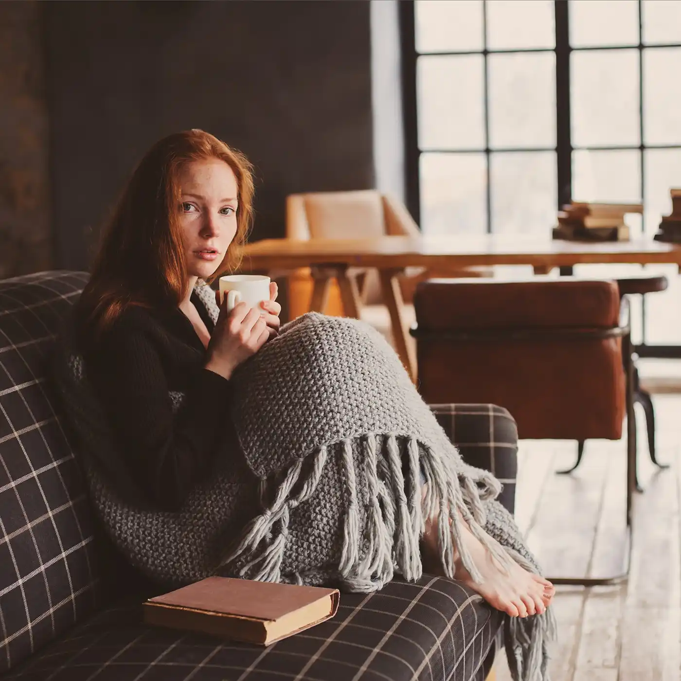 Frau sitzt eingehüllt in eine Decke mit einer Tasse auf dem Sofa – Symbolbild für Erschöpfung, innere Anspannung und psychosomatische Beschwerden.